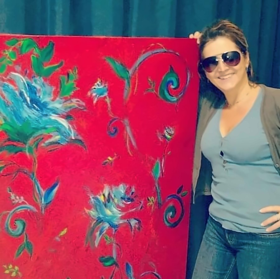 Mulher de óculos escuros sorrindo ao lado de uma tela vermelha com flores azuis estilizadas.