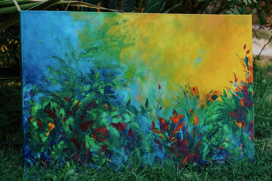 Tela em azul, verde e amarelo, com folhagem e flores vermelhas, apoiada na grama de um jardim.