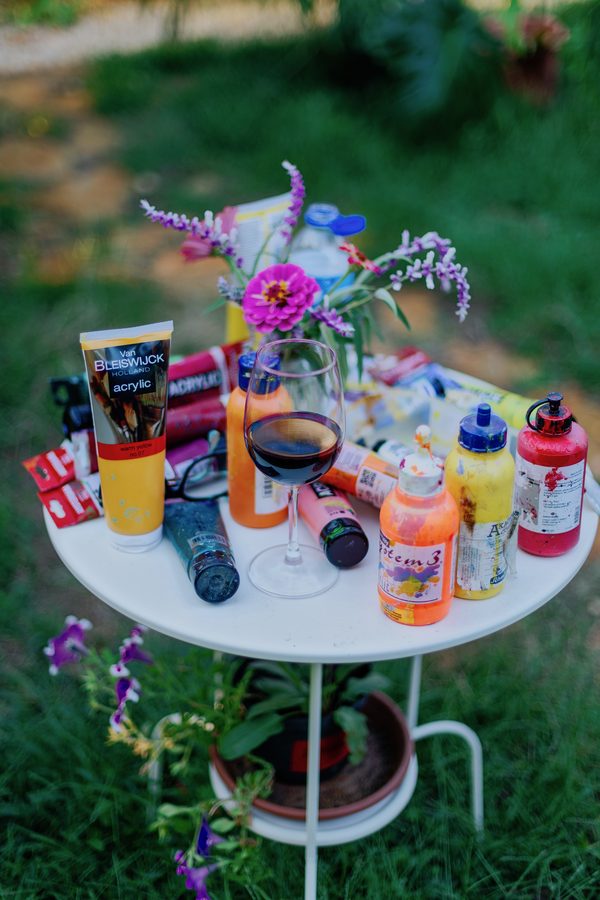 Mesa branca no jardim com tubos de tinta, vasos de flores e um copo de vinho.