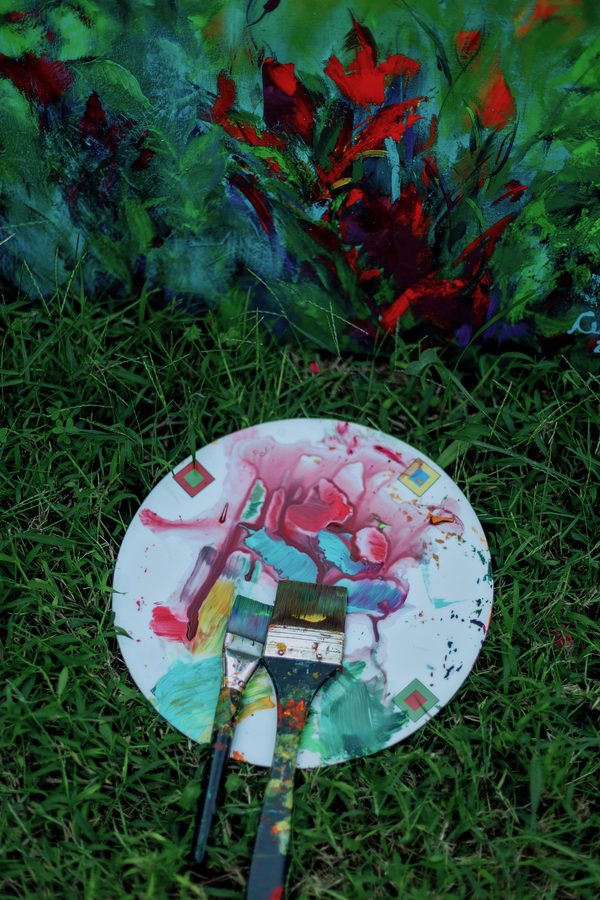 Paleta redonda com tinta rosa, azul e verde e dois pincéis, sobre a grama.