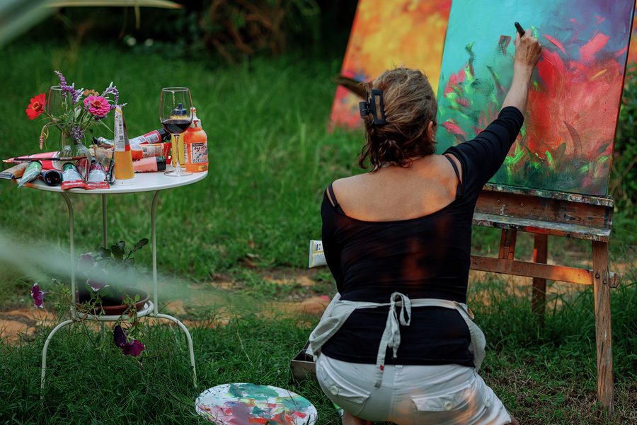 Ceres pintando ao ar livre, de costas, diante de um cavalete, com uma mesinha de tintas e flores ao lado.