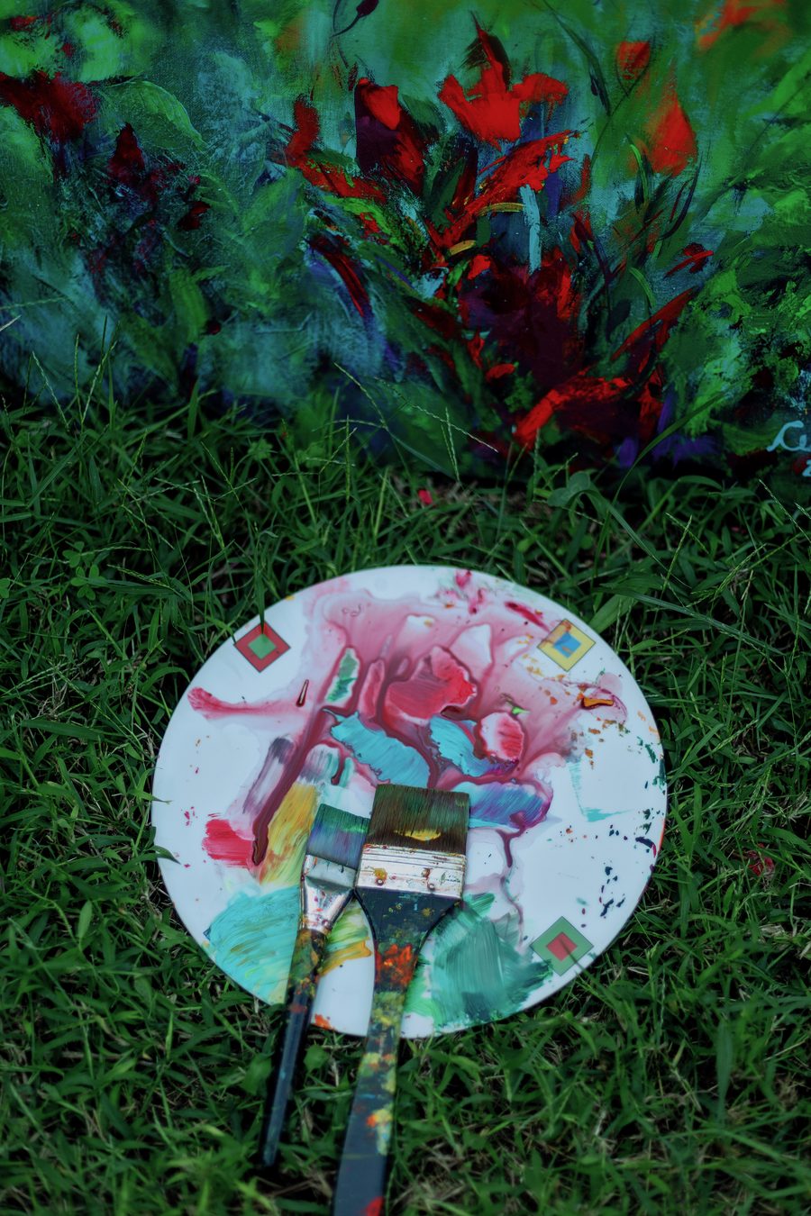 Paleta redonda com tinta rosa, azul e verde e dois pincéis, sobre a grama.
